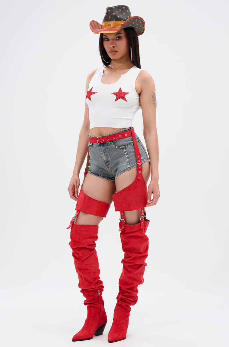 A person wears the SEEING STARS CROPPED TANK TOP—a white tank with red glitter star appliqués—paired with high-waisted denim shorts, red thigh-high boots with garter straps, and a black cowboy hat featuring gold accents.