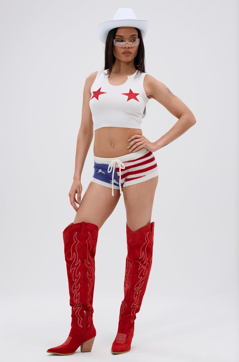A person wearing the SEEING STARS CROPPED TANK TOP, a white cowboy hat, American flag shorts in blue and red, and tall red cowboy boots stands against a plain light background.