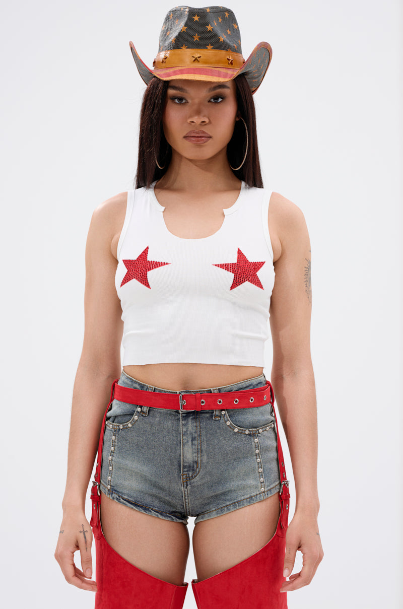 A woman models the SEEING STARS CROPPED TANK TOP with red glitter star appliqués, paired with denim shorts, a star-print cowboy hat, and red accessories, against a plain white background.