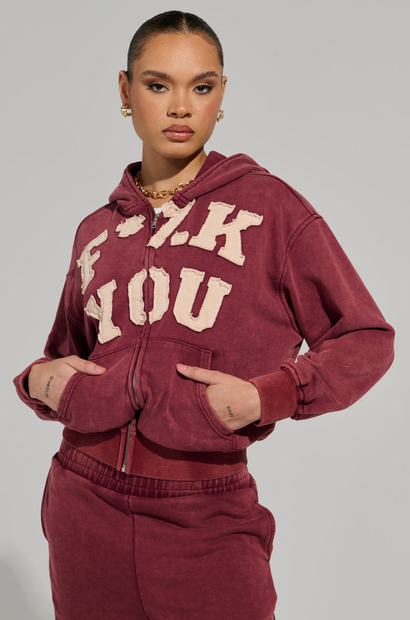 A person wearing a GRAPHIC LANGUAGE MINERAL WASH ZIP UP SWEATSHIRT IN BURGUNDY with bold lettering on the front and matching sweatpants. The background is plain and light gray. Their hair is styled neatly, complemented by earrings and a necklace.