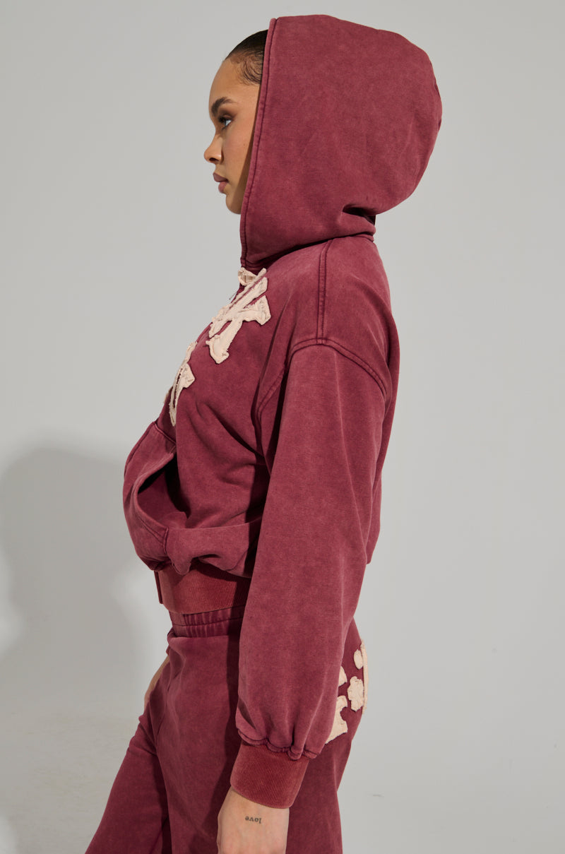 A person wearing a burgundy AKIRA Label Graphic Language Mineral Wash Zip Up Sweatshirt and matching pants stands in profile against a plain background. The sweatshirt showcases decorative text, while the pants display a sleek design on the side.