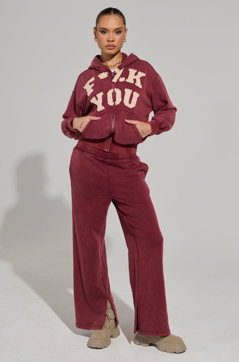 A person stands confidently wearing the GRAPHIC LANGUAGE MINERAL WASH ZIP UP SWEATSHIRT IN BURGUNDY, featuring bold lettering. The outfit is completed with wide-leg pants and beige chunky shoes, standing out against the plain light-colored background.