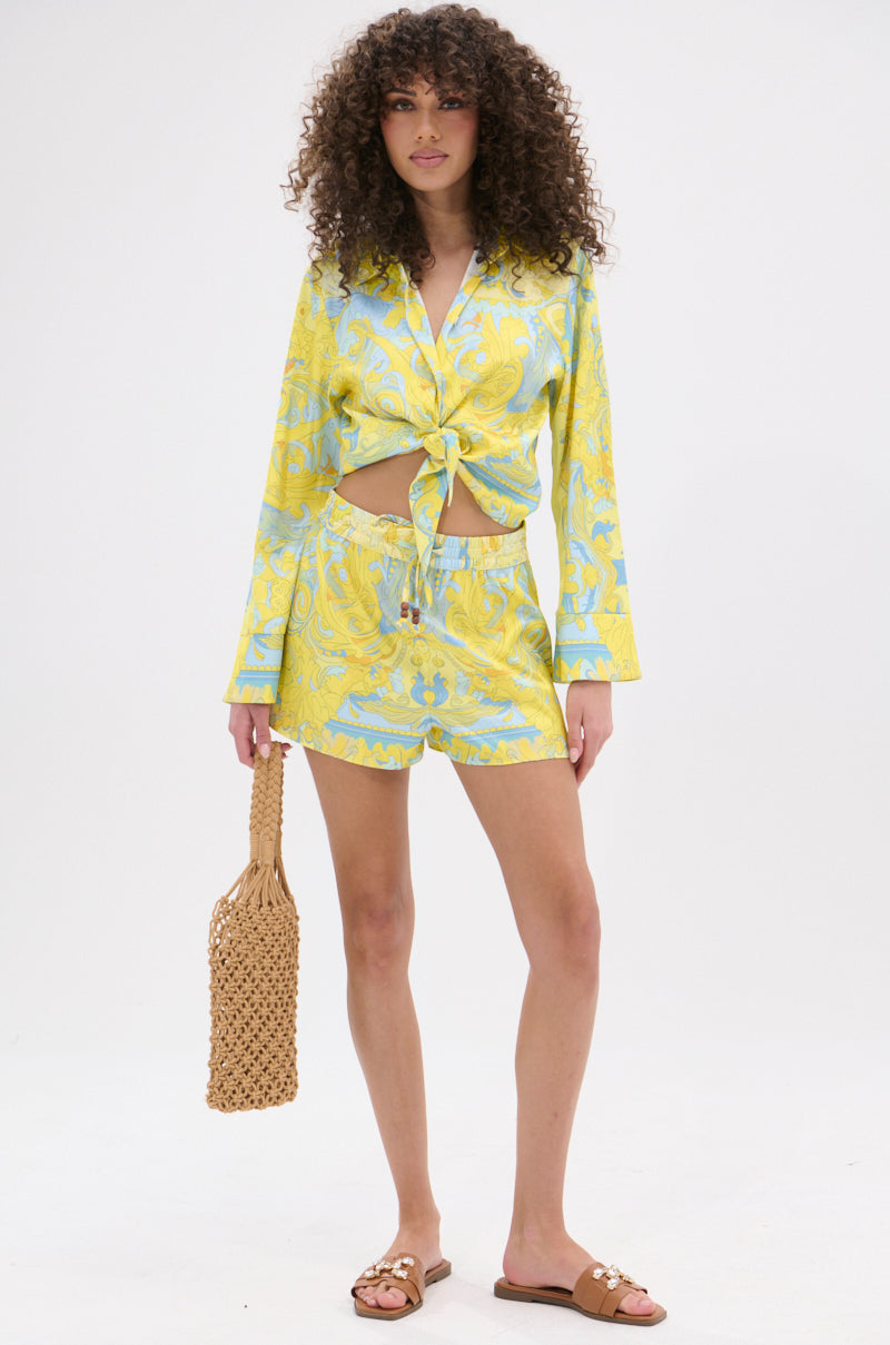 A woman with curly hair wears the FLORAL PARTY SHORT and a matching 70’s-inspired yellow and blue patterned long-sleeve shirt, tied at the waist. She holds a woven bag, wears tan slide sandals, and stands against a plain white background.