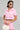 A person wearing a WORK FOR IT MINERAL WASHED CROPPED BUTTON DOWN IN PINK and a matching skirt poses confidently. They accessorize with white sunglasses, hoop earrings, a necklace, and hold a fluffy white purse with horn-like details.