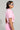 A person with short, dark hair is wearing stylish white sunglasses, gold hoop earrings, and the WORK FOR IT Mineral Washed Cropped Button Down in Pink from AKIRA Label, paired with matching high-waisted pants. They are standing sideways against a plain gray background.