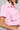 A person is wearing the AKIRA Label's WORK FOR IT Mineral Washed Cropped Button Down in Pink, featuring silver buttons and dual chest pockets. They accessorize with large hoop earrings and a beaded necklace, exuding an effortlessly chic style typical of the brand, with only their upper body visible.