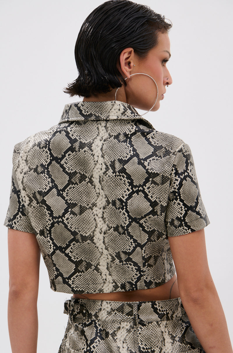 A person with short, slicked-back hair and large hoop earrings wears the VENOM CROPPED LEATHER BUTTON DOWN in a gray and black snakeskin print, shown from the back.