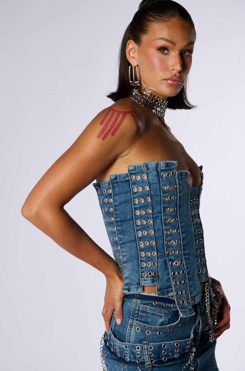 A person with sleek dark hair, pink lipstick, and large hoop earrings stands confidently with their hands on their hips, showcasing the JUST WANNA ROCK DENIM TUBE TOP— a strapless, studded denim tube top paired with stretchy patchworked denim pants. The outfit features metallic details and they have a red shoulder tattoo.