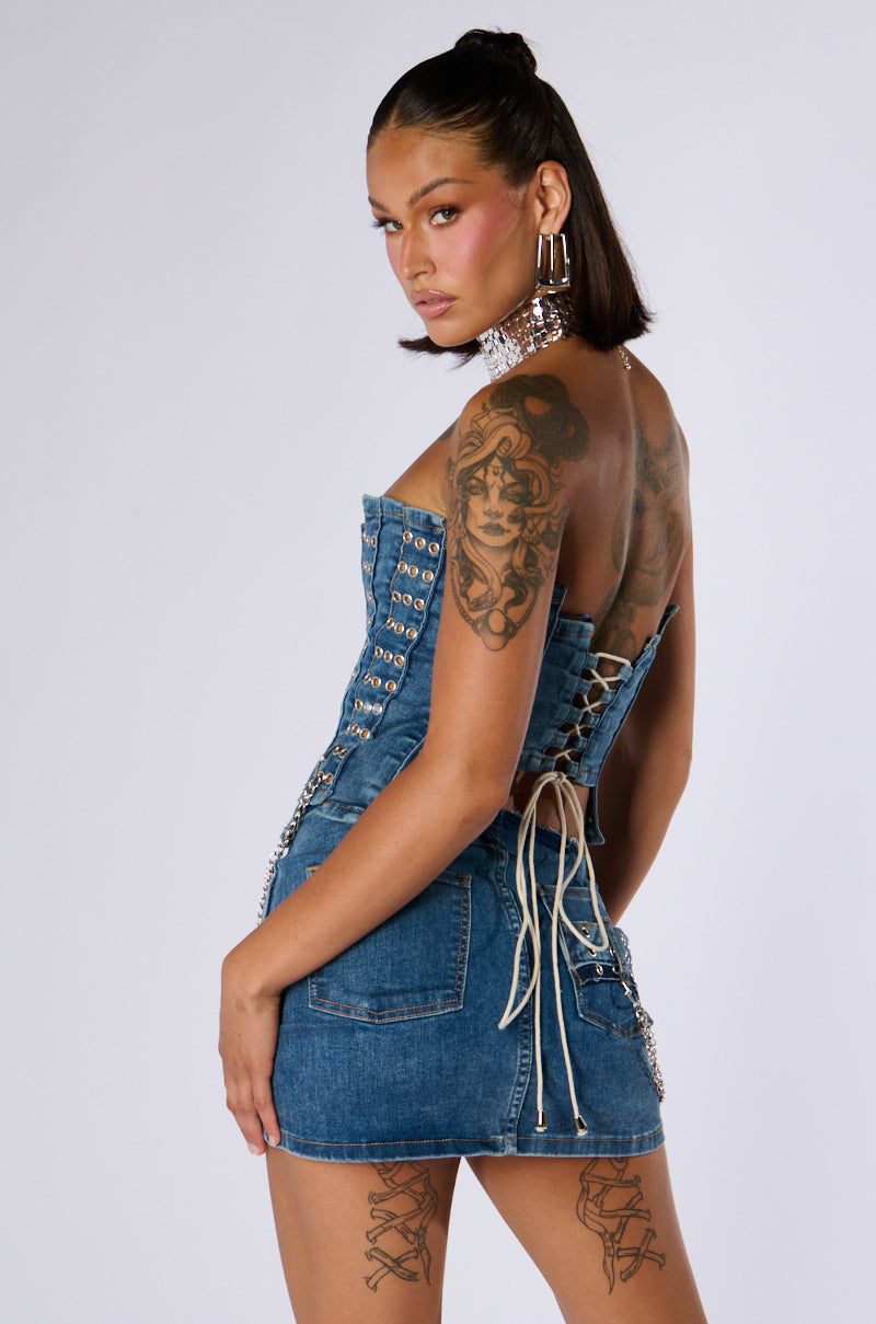 A person with long dark hair in a half-up hairstyle poses confidently. They wear the JUST WANNA ROCK DENIM TUBE TOP, a patchworked denim mini dress with lace-up details on the back, accessorized with large hoop earrings and a choker necklace. Tattoos are visible on their upper back and thighs.