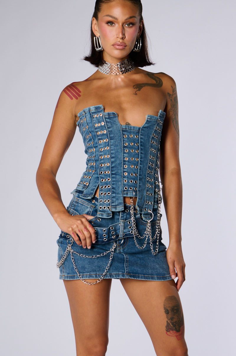 A person with long hair and tattoos is posing against a gray background. They are wearing the JUST WANNA ROCK DENIM TUBE TOP paired with a matching mini skirt adorned with chains. The outfit features eyelet and strap details in stretchy patchworked denim, completed with a choker necklace to accessorize the look.