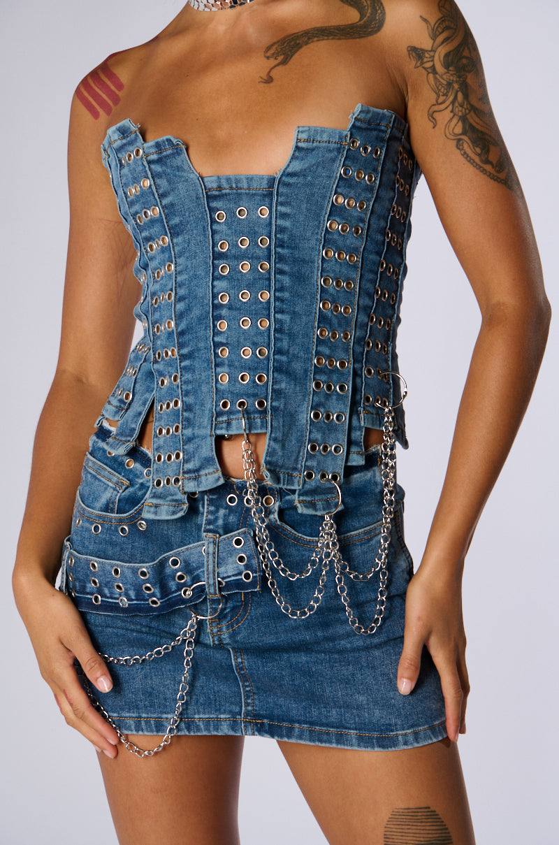 A person wearing the JUST WANNA ROCK DENIM TUBE TOP, a blue corset-inspired crop top with silver grommets and chains, paired with a matching denim mini skirt adorned with similar grommet and chain details. The person has visible tattoos on their upper arms and collarbone area.