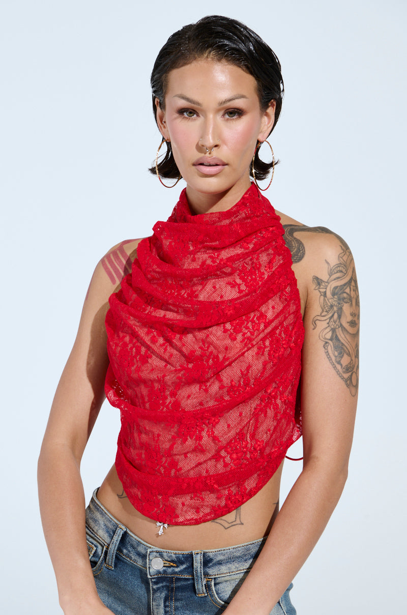 A person with slicked-back hair and large hoop earrings wears the red LETICIA LACE HALTER NECK TOP adorned with floral patterns. Sporting tattoos on their left arm, they stand against a light gray background, and blue jeans complete their stylish look.