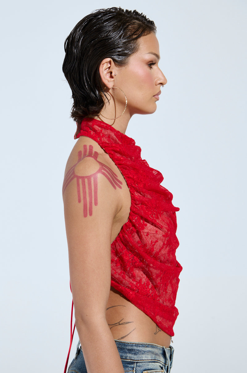 A person with short, dark, wet-look hair is wearing the LETICIA LACE HALTER NECK TOP in red and jeans. They have a circular pink design on their right shoulder and are facing left against a plain background.