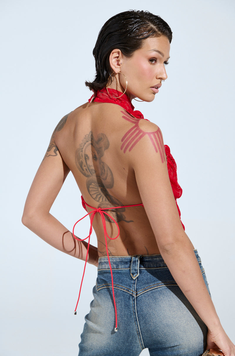 A person with short hair faces away, wearing the LETICIA LACE HALTER NECK TOP in red. They have tattoos on their upper back and arm, pair it with blue jeans, and look over their left shoulder against a light blue background.
