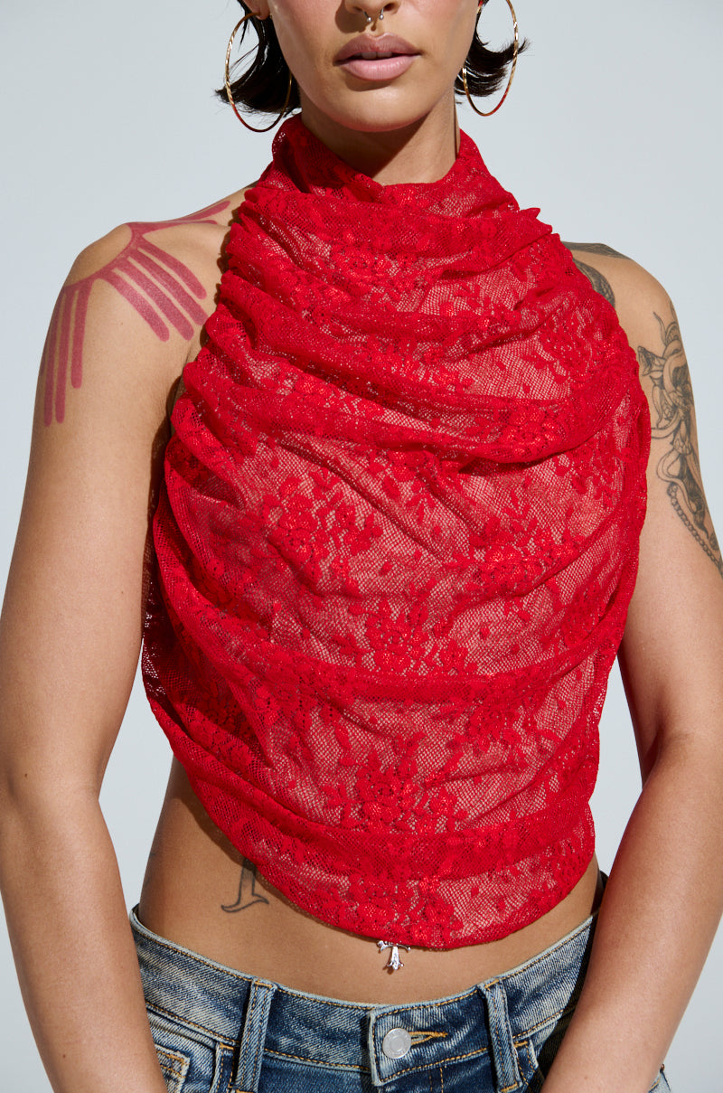 Someone wearing the LETICIA LACE HALTER NECK TOP in red, featuring a halter neck and open back, along with hoop earrings. Tattoos adorn their upper arms and chest, complemented by blue jeans. The backdrop is a plain light shade.