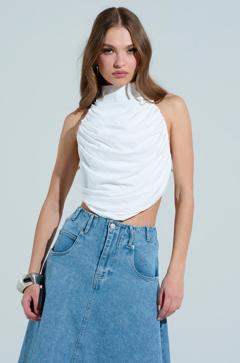 A woman with long brown hair wears a MIAMI NIGHTS HALTER NECK TIE BACK TOP and a high-waisted denim skirt. She accessorizes with a large silver bracelet and pink earrings, standing against a plain background.