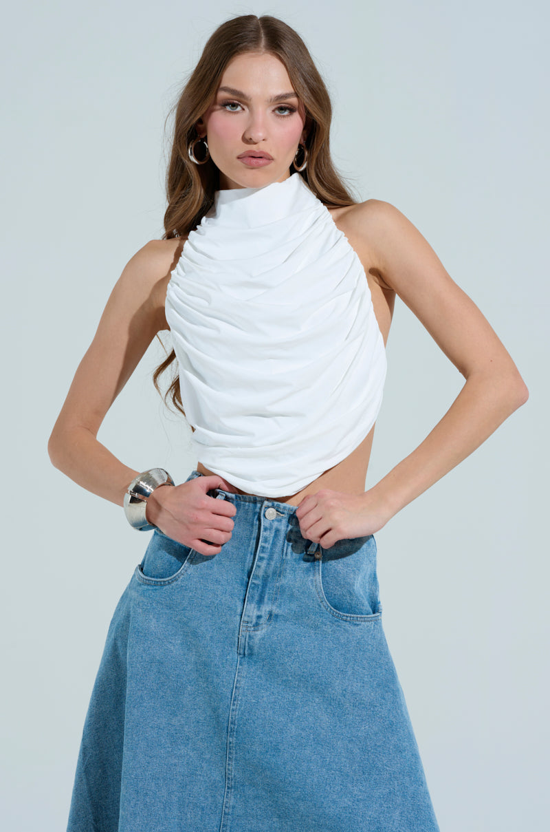 A woman poses confidently in a sleeveless MIAMI NIGHTS HALTER NECK TIE BACK TOP with ruched detailing, paired with a long denim skirt and large bracelet. Her hands rest in her skirt pockets, long hair flowing freely and hoop earrings adorning her ears against the plain background.