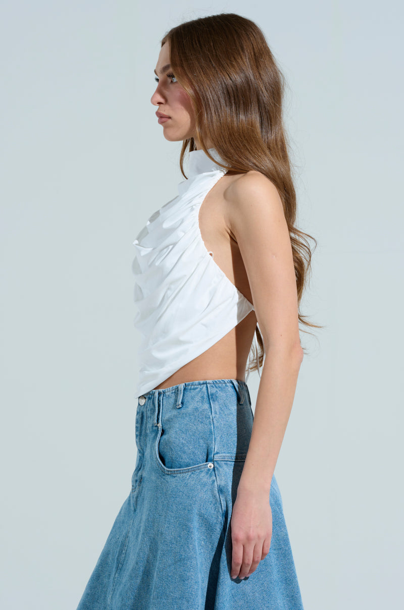 A person with long brown hair stands in profile against a plain background, wearing the MIAMI NIGHTS HALTER NECK TIE BACK TOP and a high-waisted blue denim skirt.