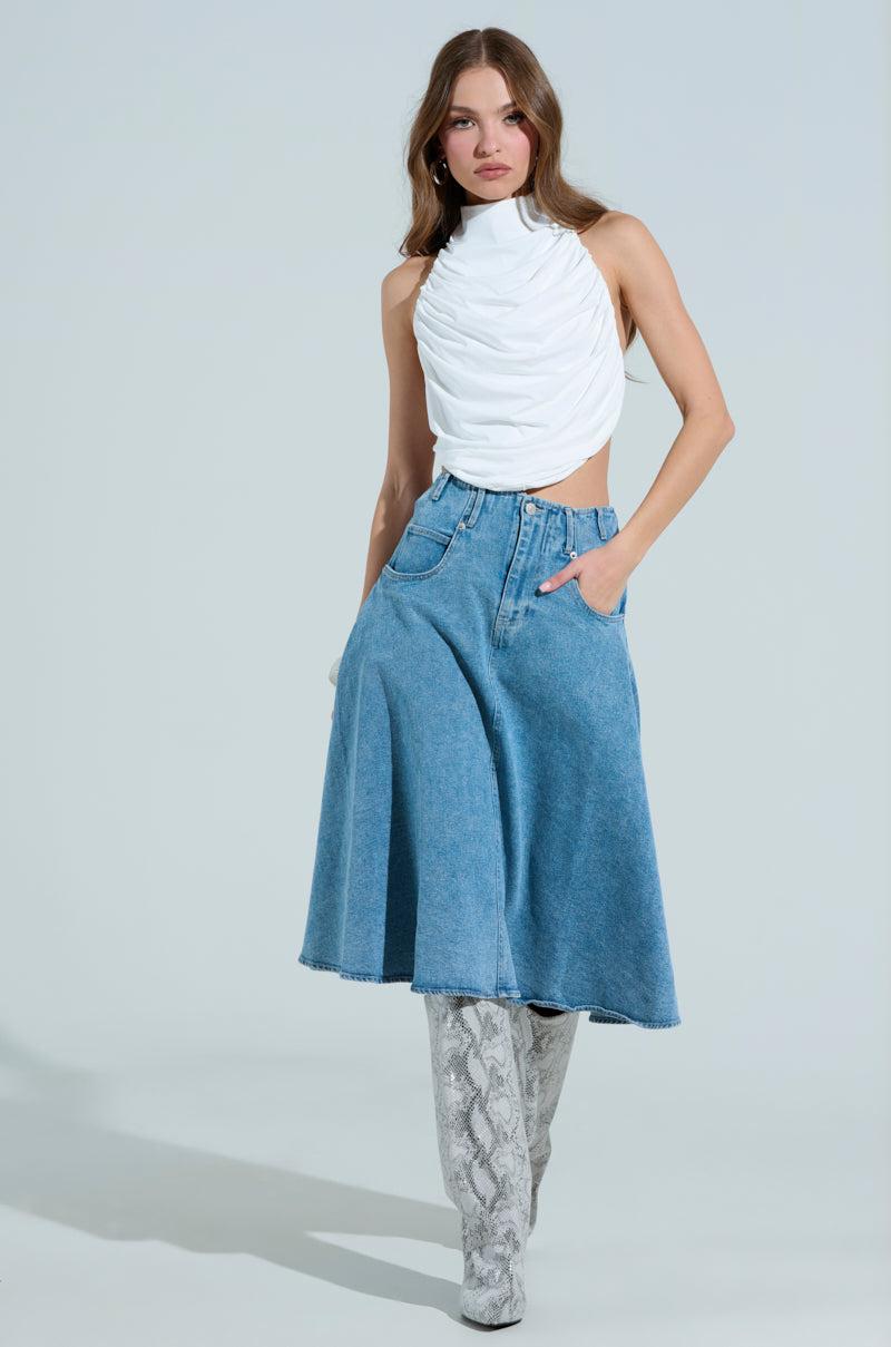 A woman confidently poses in the MIAMI NIGHTS HALTER NECK TIE BACK TOP with a blue denim skirt, styled with knee-high snake print boots and one hand in her pocket. The plain background enhances her fashionable ensemble.
