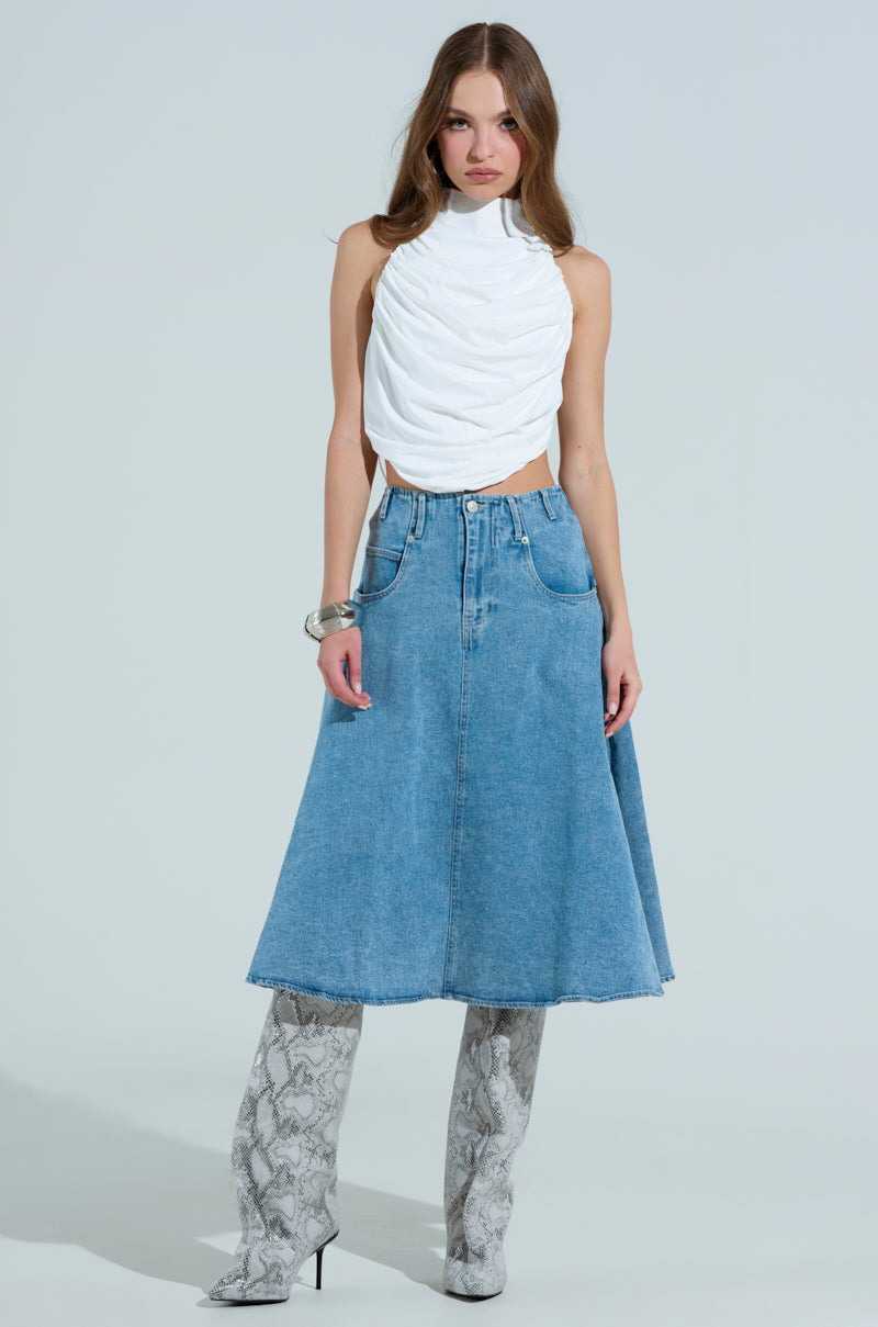 Wearing a MIAMI NIGHTS HALTER NECK TIE BACK TOP and a blue denim A-line skirt, the woman completes her look with knee-high snake-patterned boots and a large silver bracelet against a plain background.