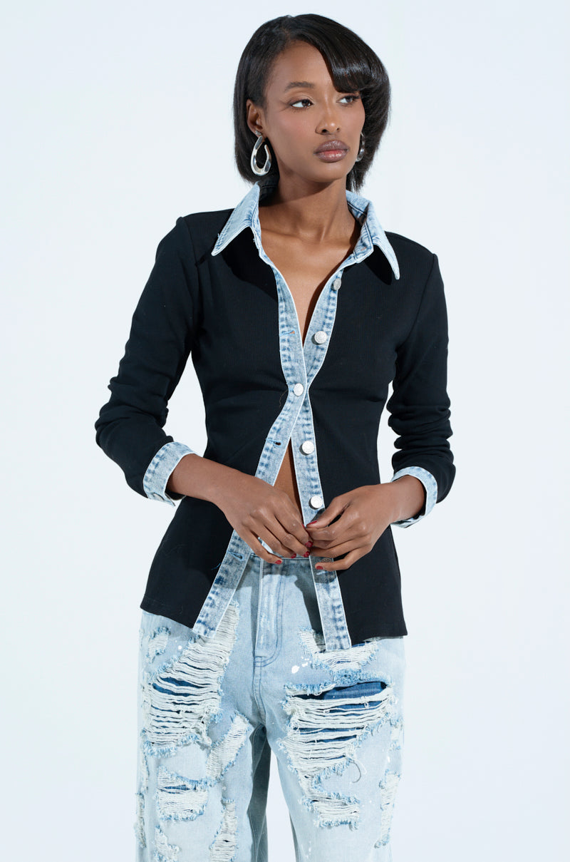 A person in a black Rib Knit Denim Trim Button Down Collared Shirt and ripped light blue jeans stands against a plain background, wearing hoop earrings and long dark hair.