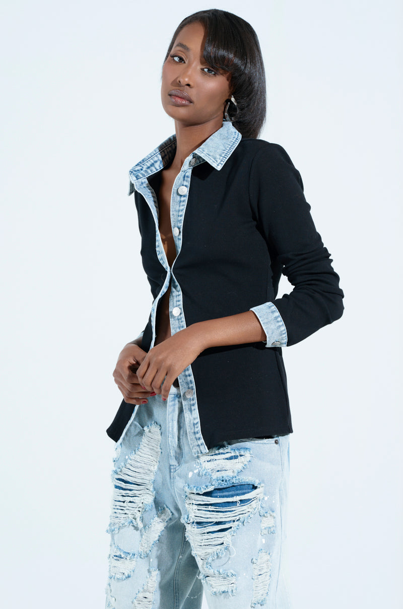A person poses confidently in a black rib knit button-down shirt with a denim trim collar and distressed blue jeans against a plain white background.