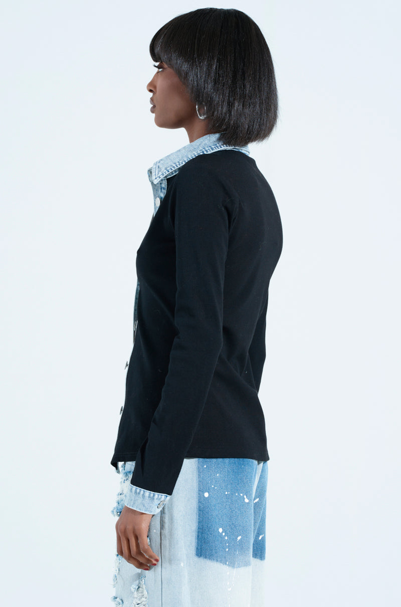 A person with short hair is standing sideways, wearing a black rib knit denim trim button down collared shirt and distressed blue jeans against a plain white backdrop.