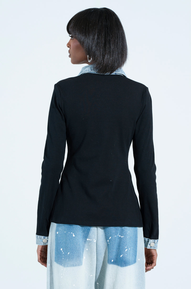 A person with shoulder-length hair is shown from behind, wearing a black blazer over a black rib knit denim trim button-down collared shirt and paint-splatter denim pants. The backdrop is plain white.