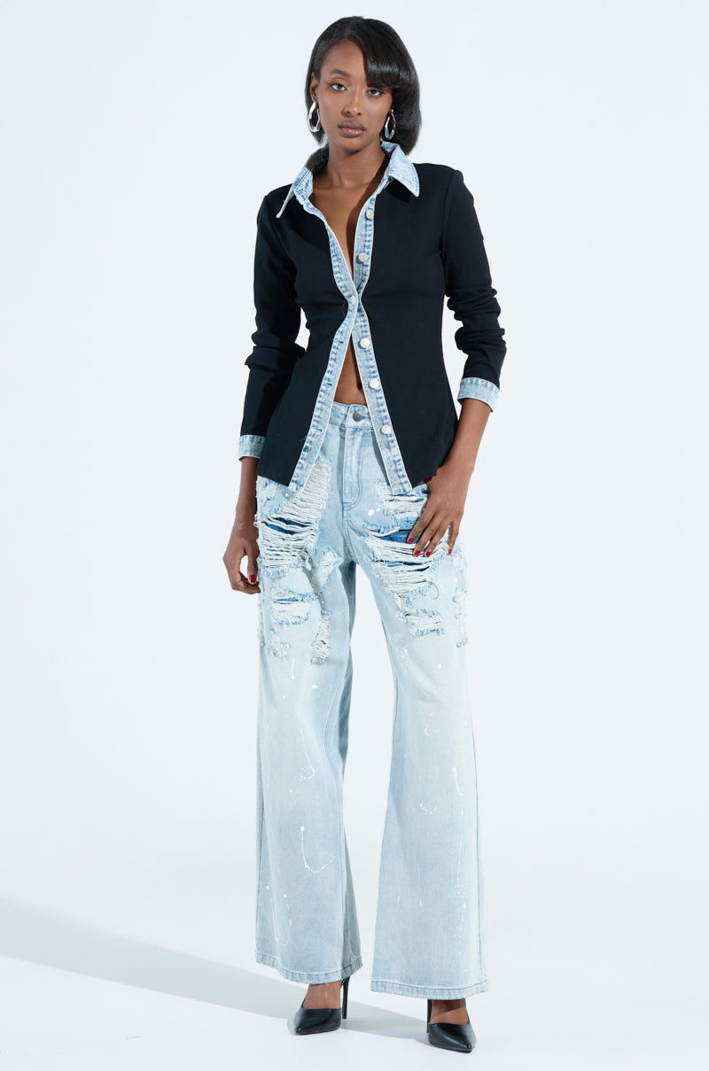 A woman exudes effortless style in a RIB KNIT DENIM TRIM BUTTON DOWN COLLARED SHIRT IN BLACK and light blue, distressed jeans. With hands tucked in her pockets and black heels, she stands confidently against a light gray background.