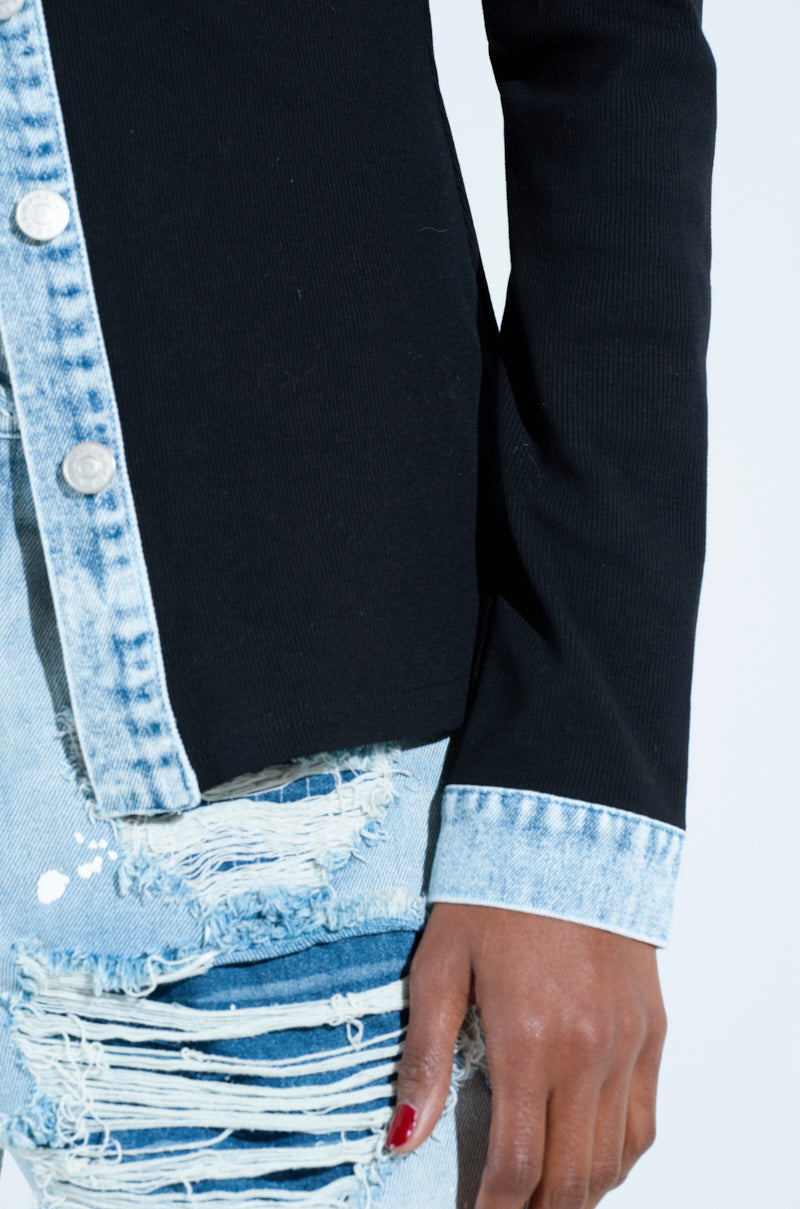 A person wears a black collared shirt with rib knit denim trim and silver buttons, paired with light blue ripped jeans. Their casual elegance is highlighted by red nail polish on one hand resting stylishly by their side.