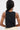 A person with long dark hair wears the RAWR TANK, a sleeveless black cropped top featuring side chains, paired with light-colored pants. The photo is taken from behind against a plain light background.