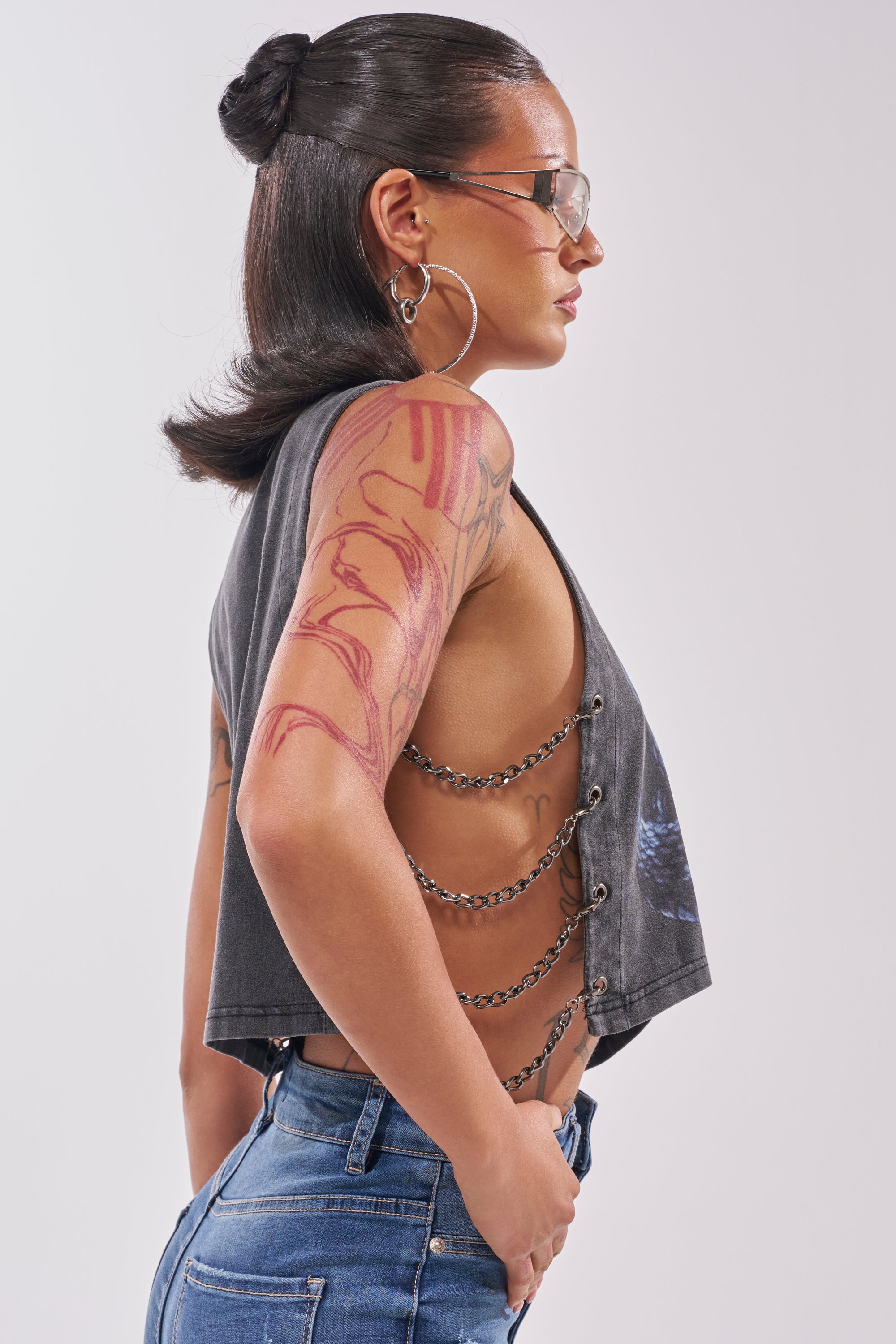A woman in profile wears sunglasses, large hoops, blue jeans, and the HISS HISS CHAIN DETAIL TANK with side chain links. Her hair is styled in a sleek bun and she has a visible tattooed arm. 3