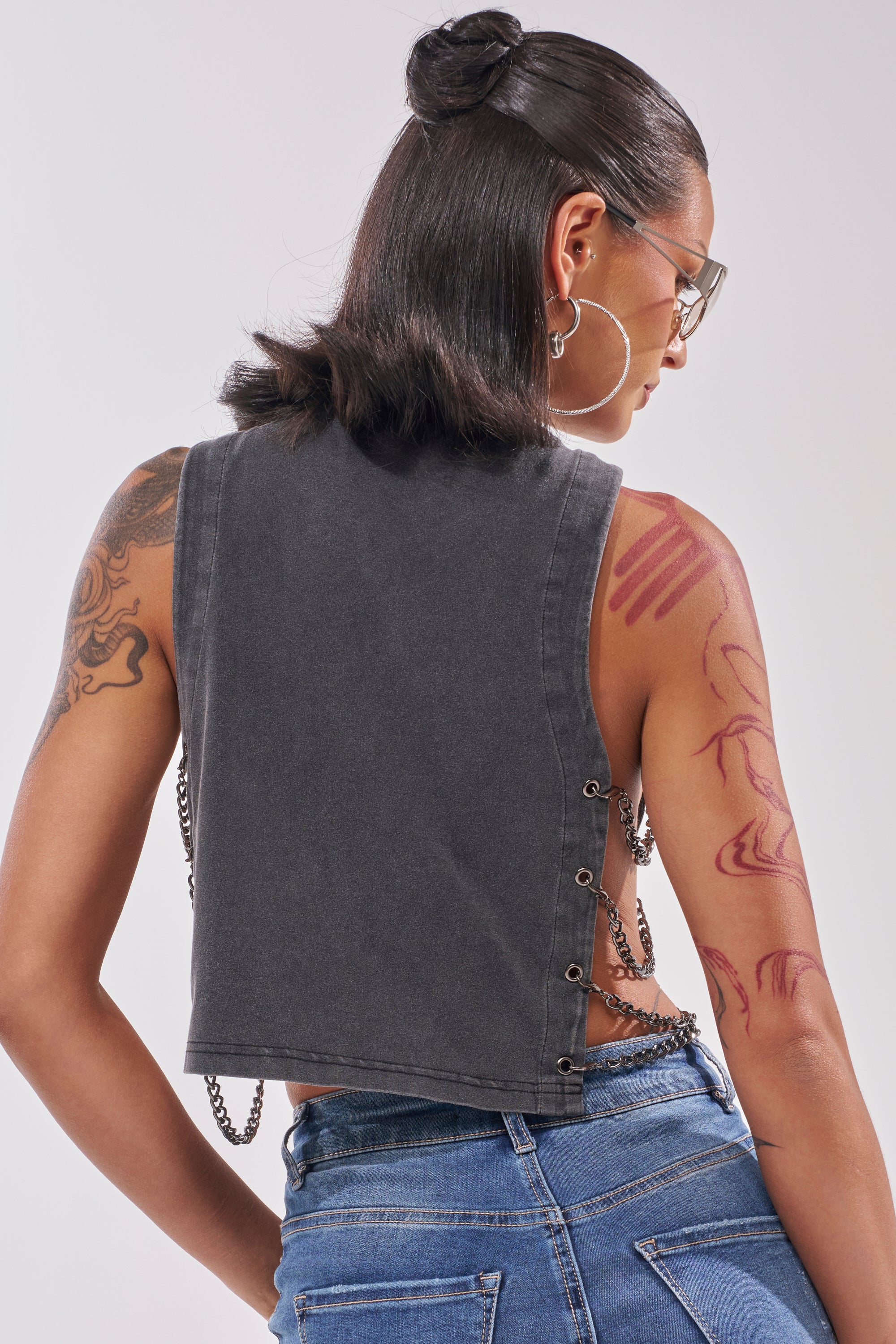 A person with long dark hair in a half-up bun is seen from the back wearing glasses, hoop earrings, blue jeans, and the HISS HISS CHAIN DETAIL TANK. Tattoos are visible on their right shoulder and arm. 4