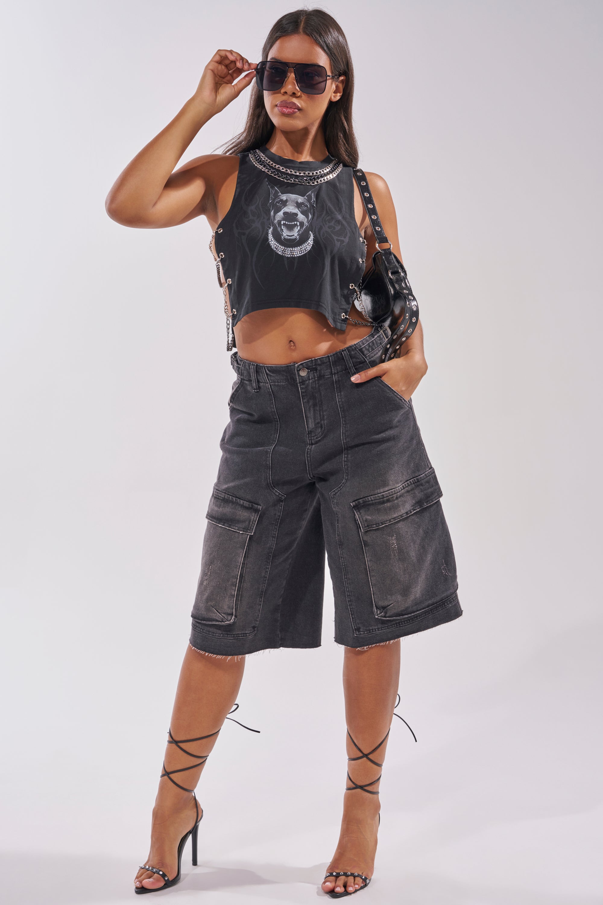 A woman in the GOT THAT DOG IN ME EMBELLISHED TANK and long black denim shorts with large pockets and chains stands confidently in strappy heels, one hand in her pocket and the other adjusting her sunglasses. 9