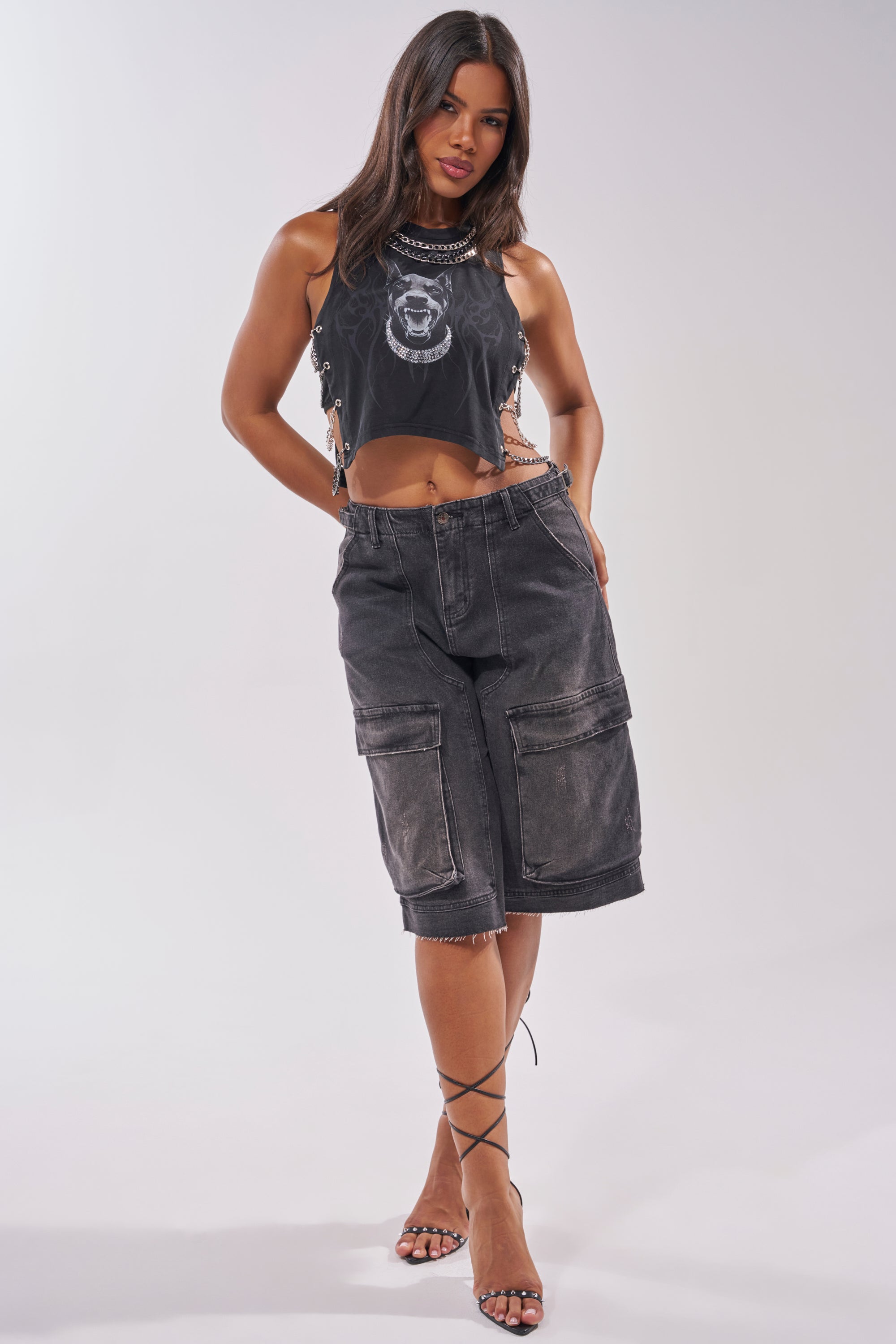 A woman models the GOT THAT DOG IN ME EMBELLISHED TANK—sleeveless, black, with graphic print and lace-up sides—styled with long black cargo shorts and strappy high-heeled sandals. 10