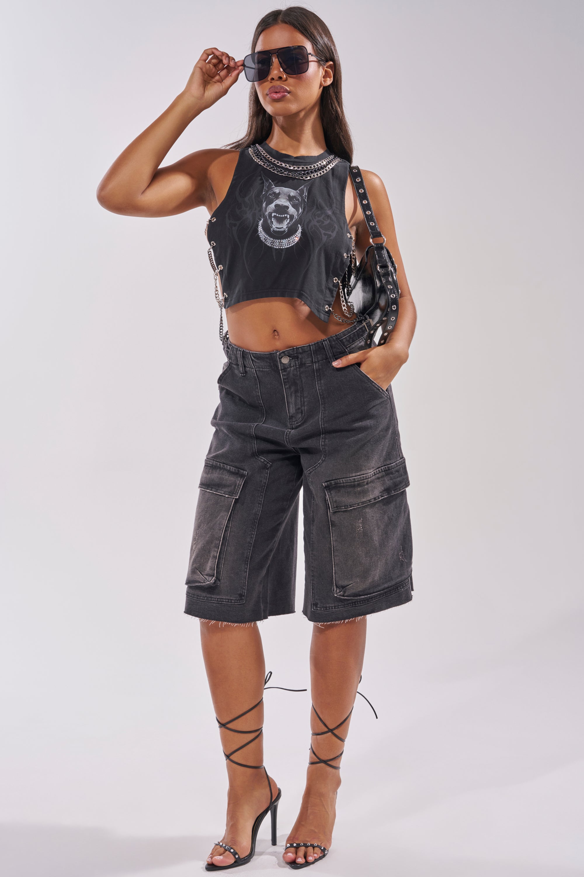 A woman poses in the GOT THAT DOG IN ME EMBELLISHED TANK—styled with long black cargo shorts, strappy heels, sunglasses, and a studded bag—showcasing statement streetwear against a plain light background. 8