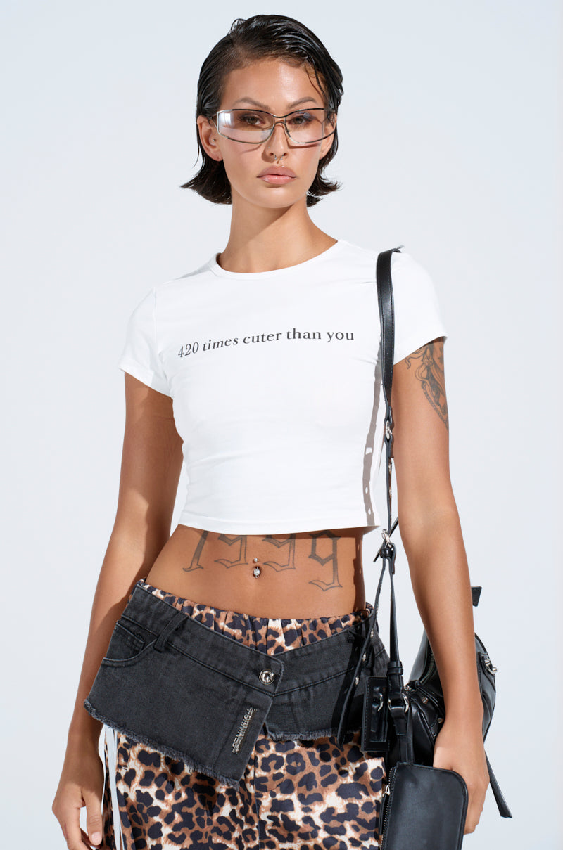 A person with short dark hair wears trendy sunglasses, a 420 TIMES CUTER THAN YOU BABY TEE, leopard print pants, and a denim accessory. They display an AKIRA Label black bag while posing against a light background. 0