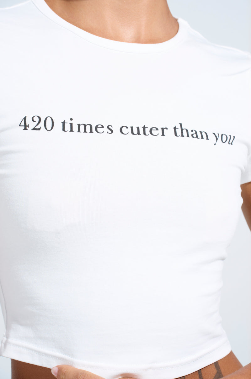 A close-up showcases an AKIRA Label baby tee named "420 TIMES CUTER THAN YOU BABY TEE." The white crop top features the playful phrase against a plain, light backdrop, highlighting its cheeky message. 5