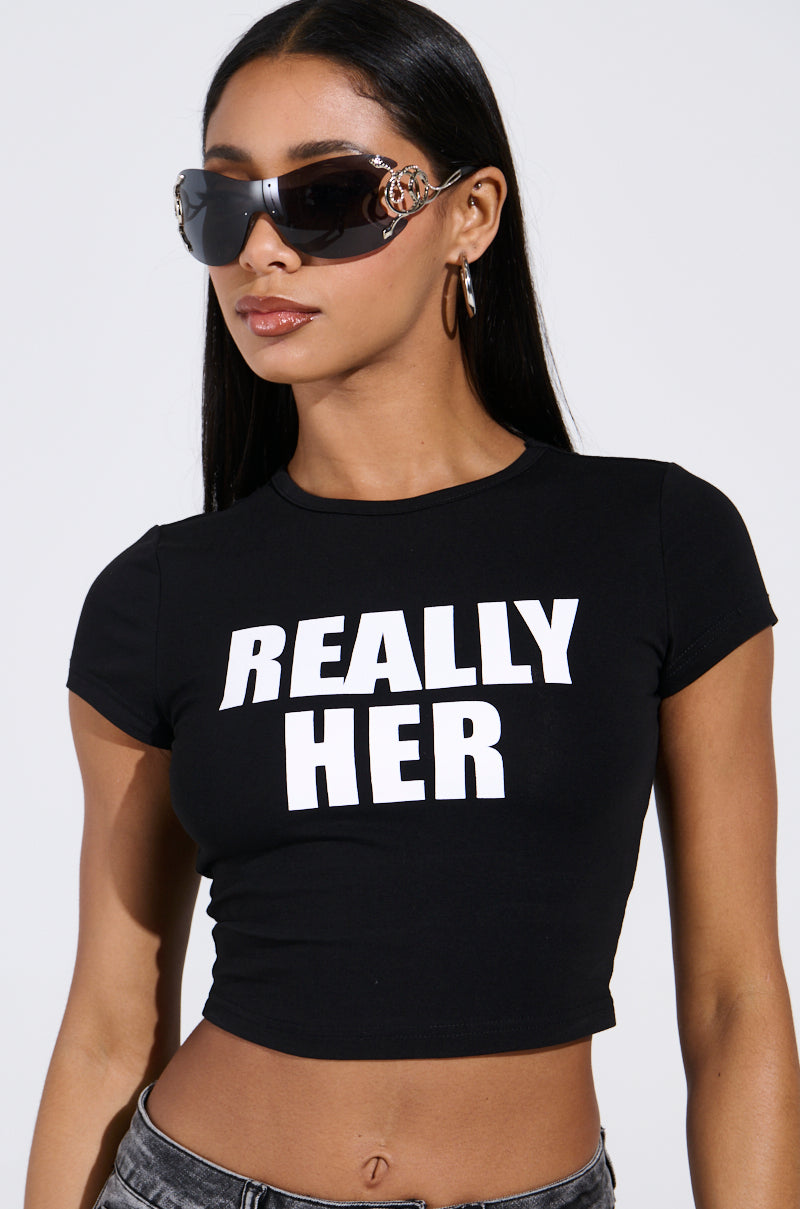 A person with long dark hair wears oversized sunglasses and a black I'M REALLY HER BABY TEE by AKIRA Label, featuring "REALLY HER" in bold white letters. They complement it with hoop earrings and jeans for a chic silhouette against a plain background.