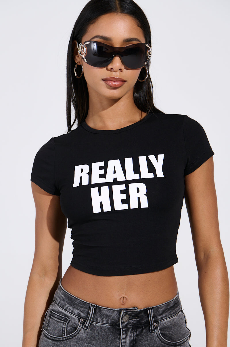 A woman poses confidently against a plain backdrop, wearing dark sunglasses and an I'M REALLY HER BABY TEE with a cropped silhouette. Her long, straight hair cascades down as she strikes her pose.