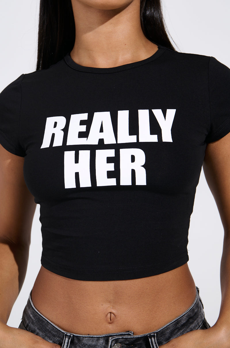 Someone is wearing the black "I'M REALLY HER BABY TEE" featuring bold white text. The image highlights the cropped fit, showing their arms and midriff, paired with gray jeans from the AKIRA Label.