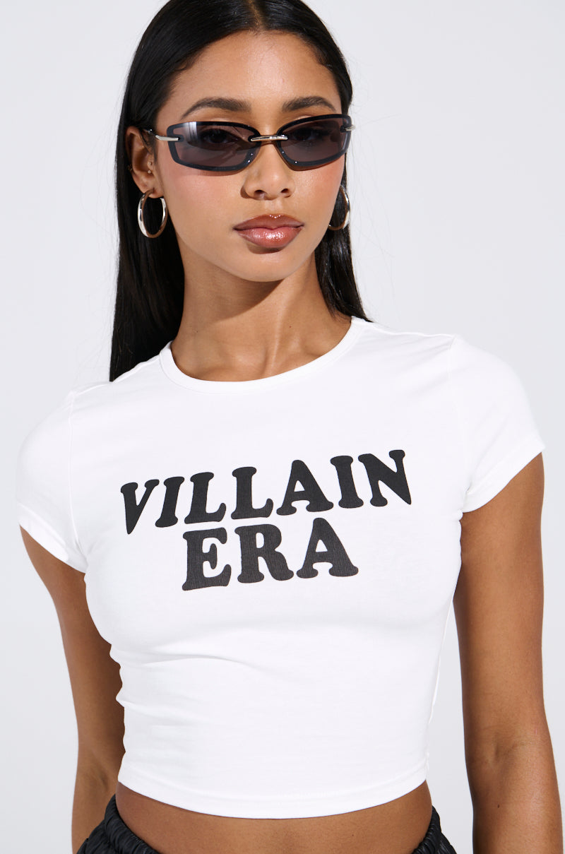 A person in sunglasses, hoop earrings, and a VILLAIN ERA TEE made from stretchy cotton. The white crop top features a chic cropped design, perfectly complementing their long dark hair against a plain backdrop.