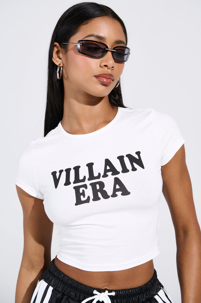 A woman with long dark hair wears rectangular sunglasses, hoop earrings, and the sleek cotton Villain Era Tee. Her outfit includes a white crop top with bold black letters and striped pants, set against a plain light gray background.