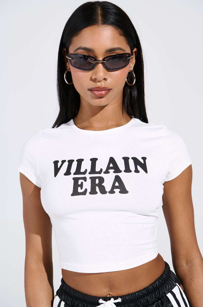Sporting long dark hair and sunglasses, this style icon rocks hoop earrings and a cropped white "VILLAIN ERA TEE," perfectly complemented by black and white striped pants. The outfit looks effortlessly cool against a plain backdrop, exuding confidence.
