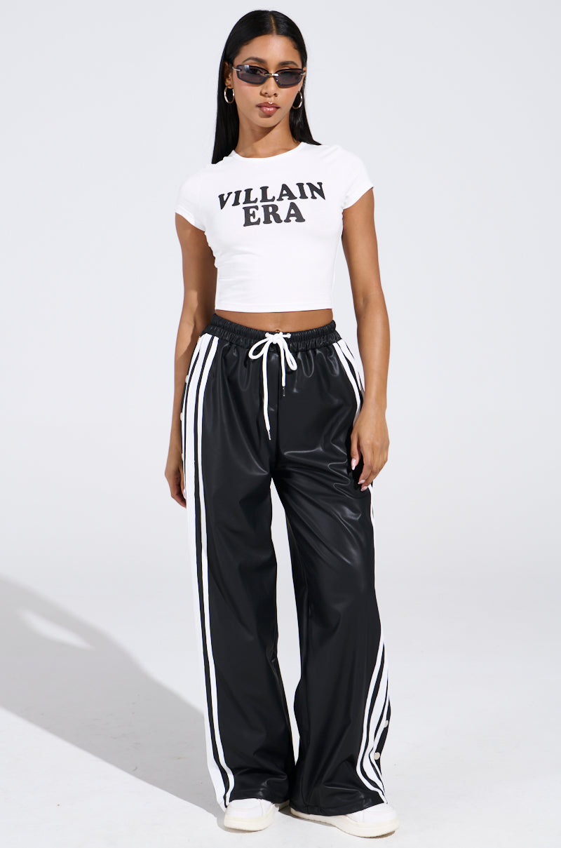 A woman in sunglasses poses against a plain backdrop, her VILLAIN ERA TEE featuring a cropped silhouette. She pairs it with black track pants sporting white stripes and finishes the look with white sneakers, exuding effortless coolness in stretchy cotton attire.