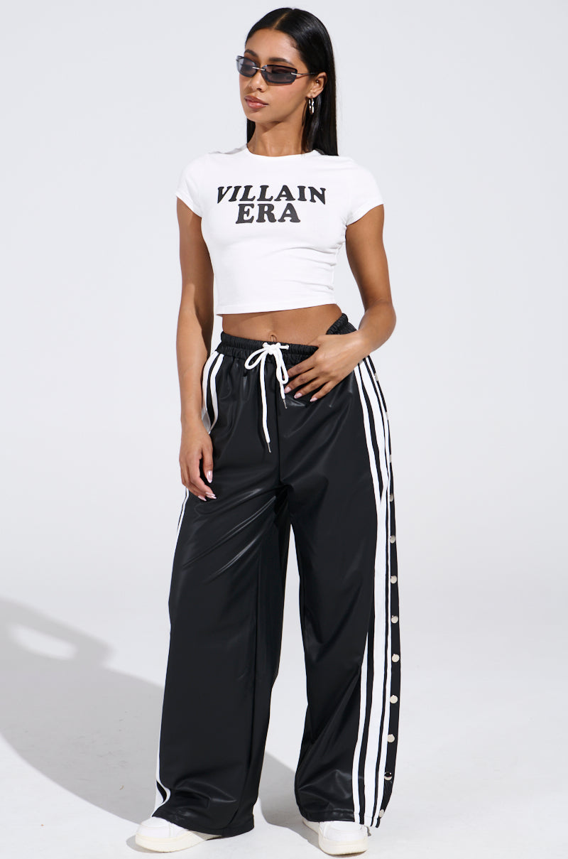 A woman poses confidently against a plain backdrop in a stretchy cotton VILLAIN ERA TEE, featuring a cropped silhouette. She pairs it with black track pants with white stripes, white sneakers, and chic sunglasses, radiating effortless style.