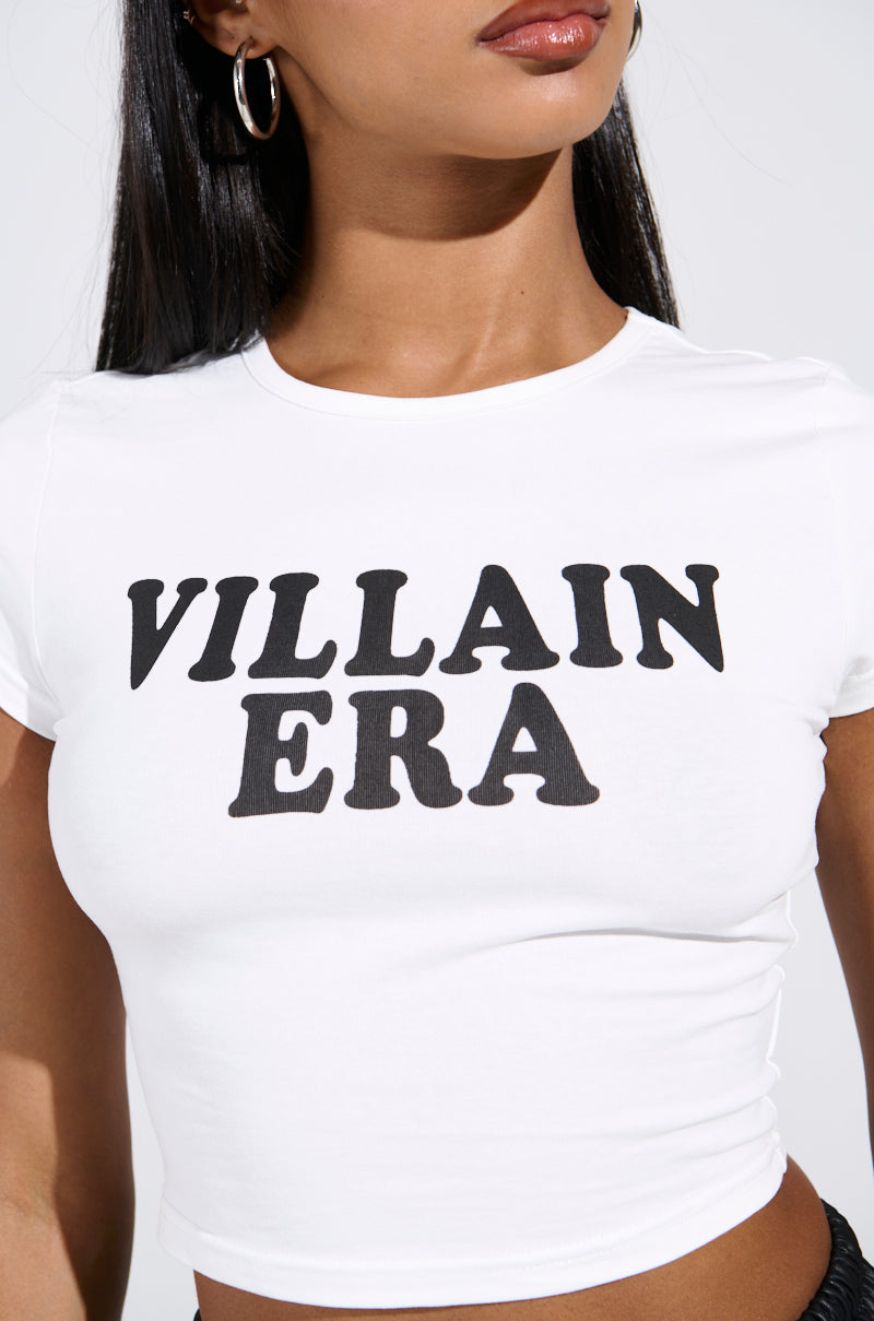 A person with long dark hair and hoop earrings wears a stretchy cotton VILLAIN ERA TEE featuring bold black letters on white fabric. The cropped silhouette stands out against a plain white background.