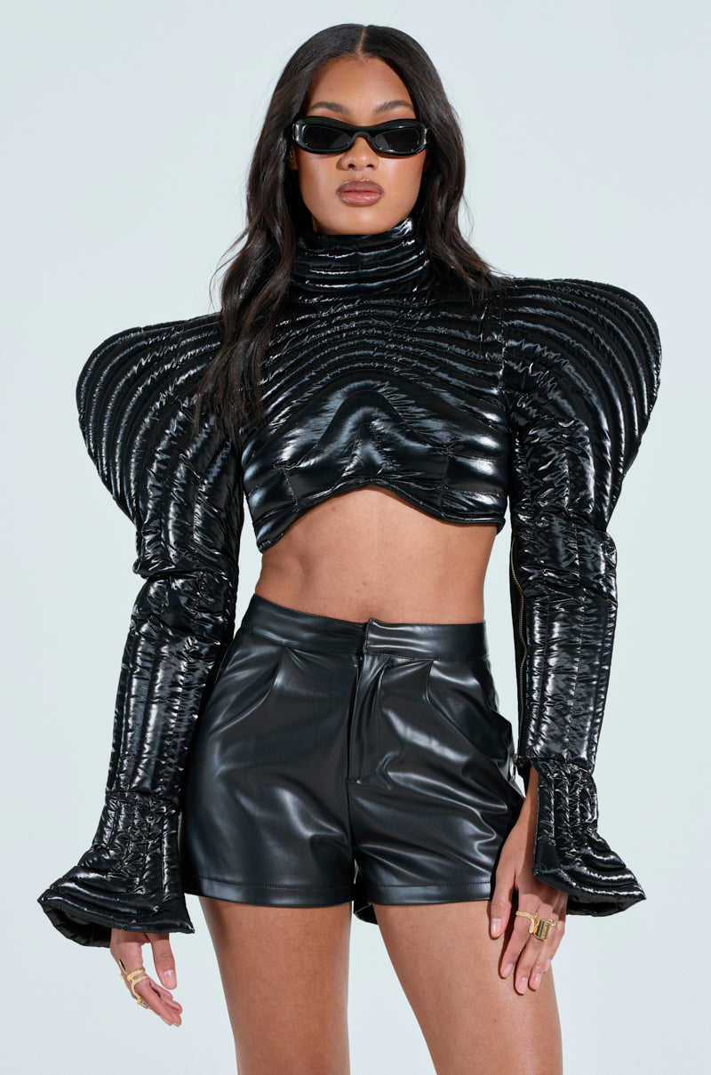 A person confidently wears the KEEP IT COMING TOP, an ultra-cropped, futuristic outfit with exaggerated shoulders and flared sleeves. They pair it with black sunglasses and have long, wavy hair against a plain, neutral background.