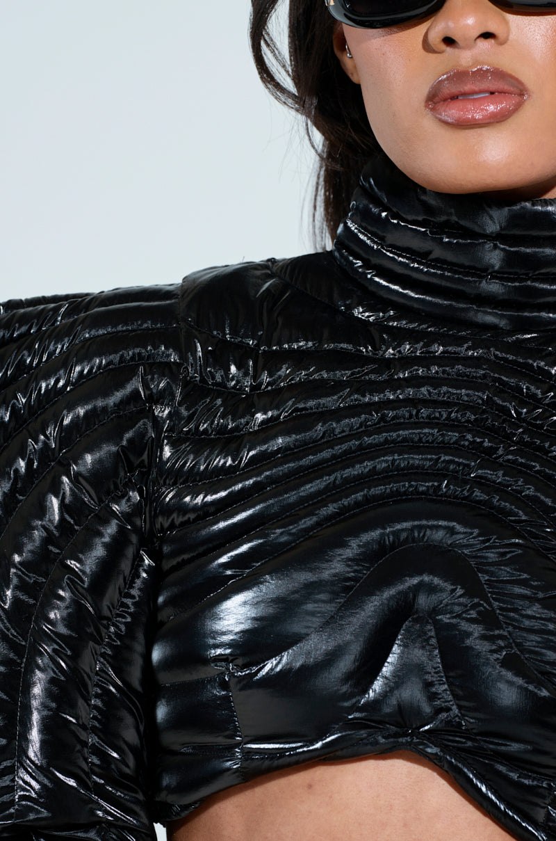 Sporting the ultra-chic KEEP IT COMING TOP, a person dons a shiny black jacket with intricate stitching and high collar, paired with large sunglasses. The image focuses on the paneled puffer design and glossy fabric, accentuating their striking style in an ultra-cropped fit.  .