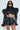 A person confidently wears the KEEP IT COMING TOP, an ultra-cropped, futuristic outfit with exaggerated shoulders and flared sleeves. They pair it with black sunglasses and have long, wavy hair against a plain, neutral background.